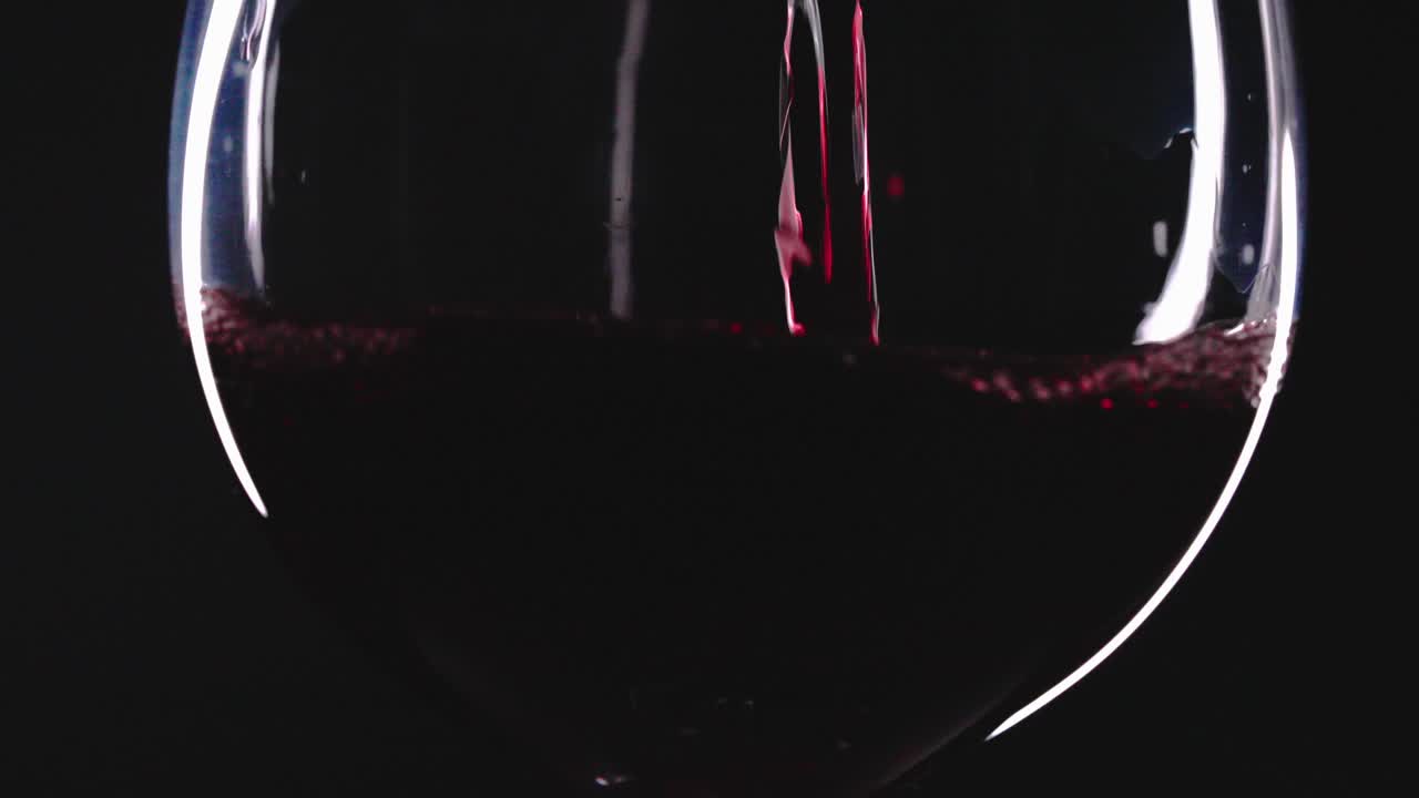 Slow-motion video of red wine being poured into a glass, highlighting its deep colour, swirling motion, and elegant flow against a dark background. (4K)