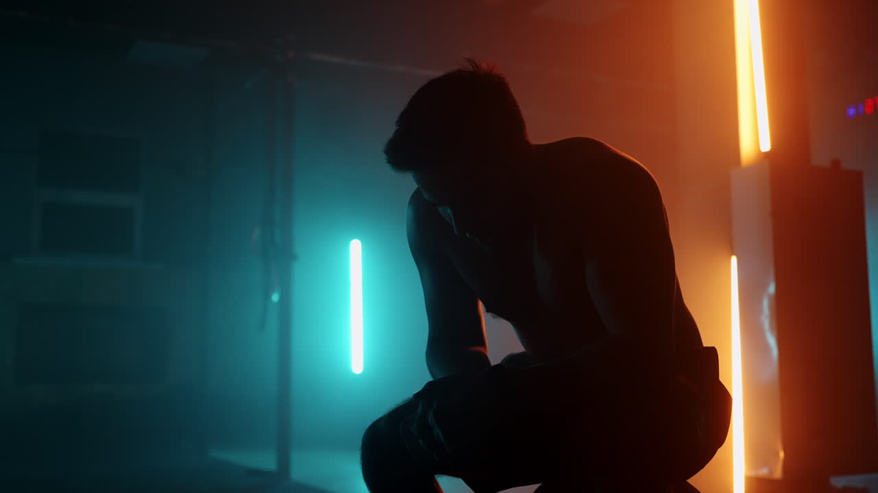 疲れたアスリートは ⁇ スローモーションの運動を実行した後 ⁇ ネオン照明の下でバーベルに座っている間 ⁇ 重く息を吸って休息しています ⁇ 回復し ⁇ 新しいブレークスルーの準備をします ⁇