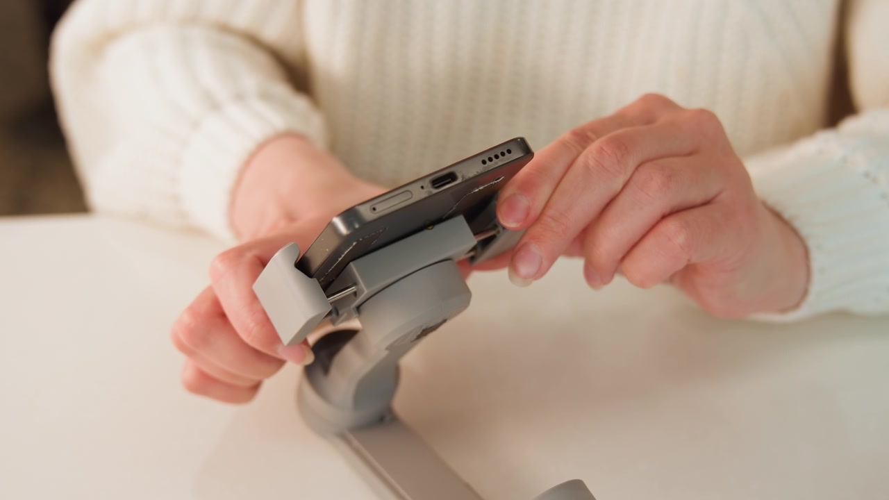 Close frontal view of hands in white sweater manipulating small printed circuit board mounted in plastic tabletop clamp, with fingers aligning components, checking placement, then pausing over tidy
