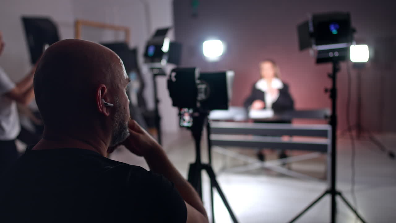 Bald bearded cameraman works backstage in photo studio. Modern professional equipment recording a video with a woman sitting at desk at blurred backdrop.