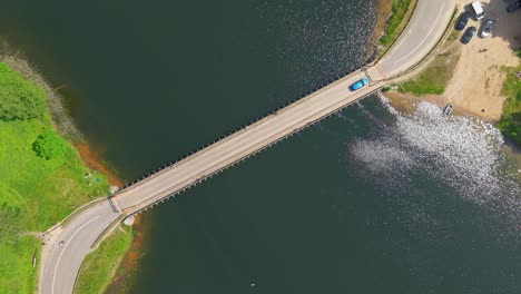 Aerial top down view of a blue car driving over a bridge crossing a river with green fields and parked cars on a sunny summer day
