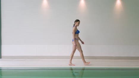 Fit-woman-in-bikini-walks-along-edge-of-indoor-swimming-pool