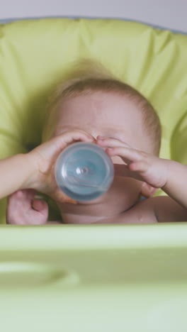 careful blonde girl gives bottle of water to unhappy little boy sitting in soft highchair in light room