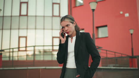 White girl in professional outfit talking on phone with tired expression in urban area, hand in pocket, red and glass building in background, showing signs of fatigue and emotional exhaustion
