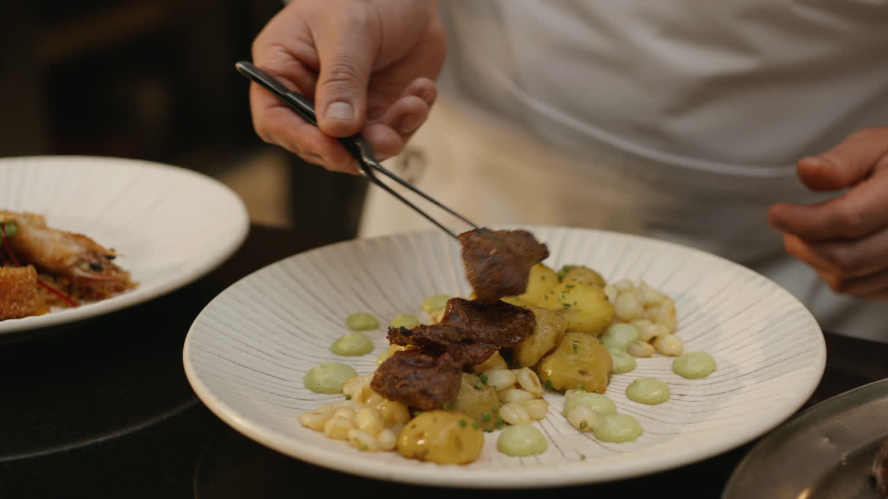 A chef uses tongs to carefully place pieces of meat on a bed of potatoes and corn