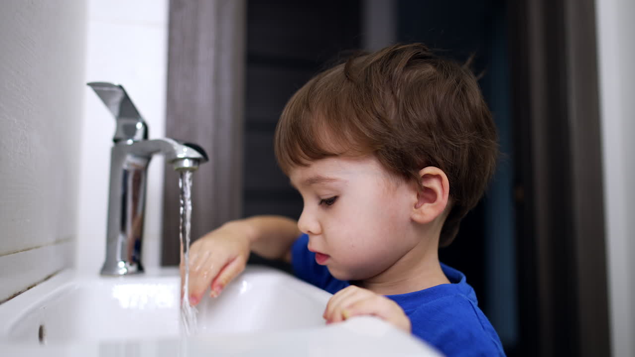 Lovely cute toddler standing at the sink in the bathroom. Caucasian boy opens and closes the tap. Close up.