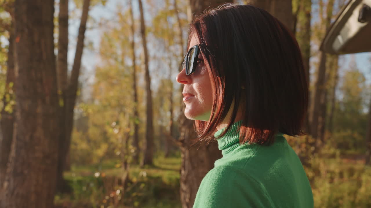 Donna in campeggio nella foresta illuminata dal sole che indossa occhiali da sole, profilo con maglione verde in riva al lago, bagagliaio dell'auto visibile, luce dell'ora d'oro, fogliame autunnale, sguardo contemplativo, campeggiatrice rilassata che si gode la natura.