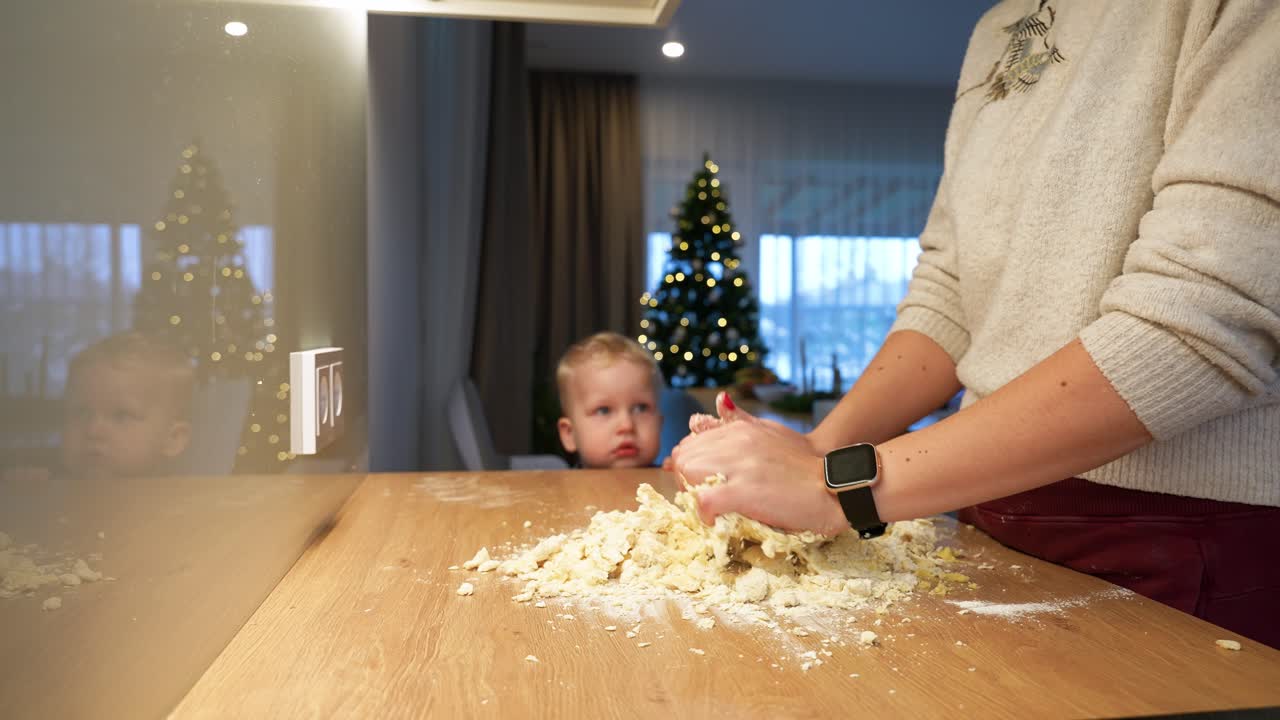 A cozy indoor scene features a person kneading dough on a wooden table while a child watches. Warm ambient lighting enhances the homely atmosphere, with a decorated Christmas tree in the background