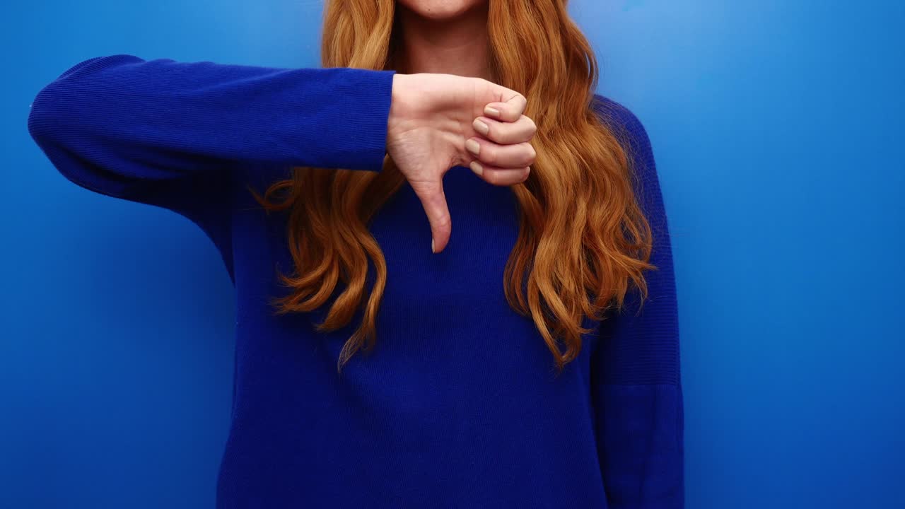 Young redhead woman in blue shirt showing thumbs down gesture, isolated on the blue background