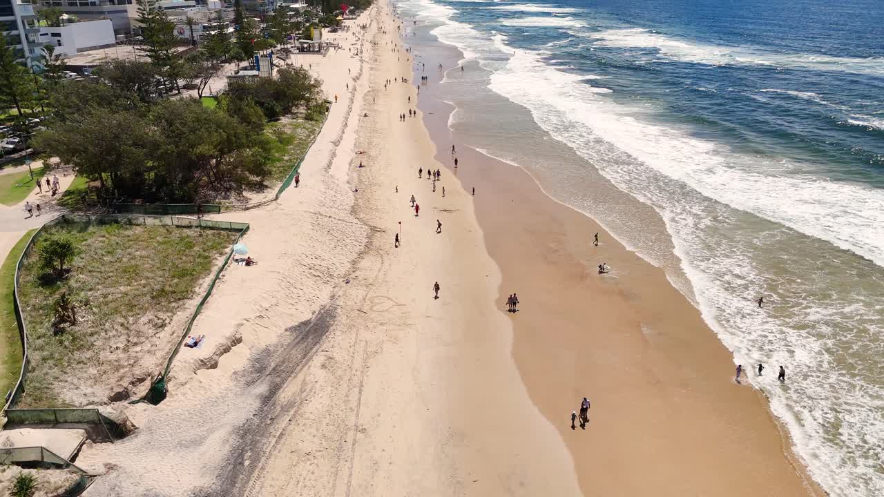 Aerial footage captures people strolling along Broadbeach, Gold Coast. Sunny day, clear skies, and gentle ocean waves