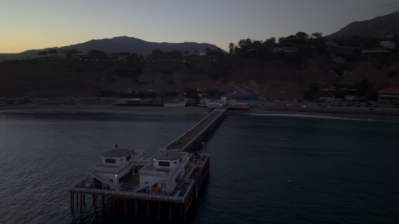 Sunset view of Malibu Pier, California, showing surfers near the lagoon outlet with Pacific Coast Highway and the coastal mountains behind. Soft dusk colors reflect across the water and shoreline