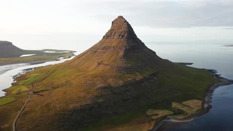 Kirkjufell mountain in Iceland, rising sharply near a serene coastline, surrounded by calm waters, green fields, and breathtaking natural landscapes, creating a stunning iconic view