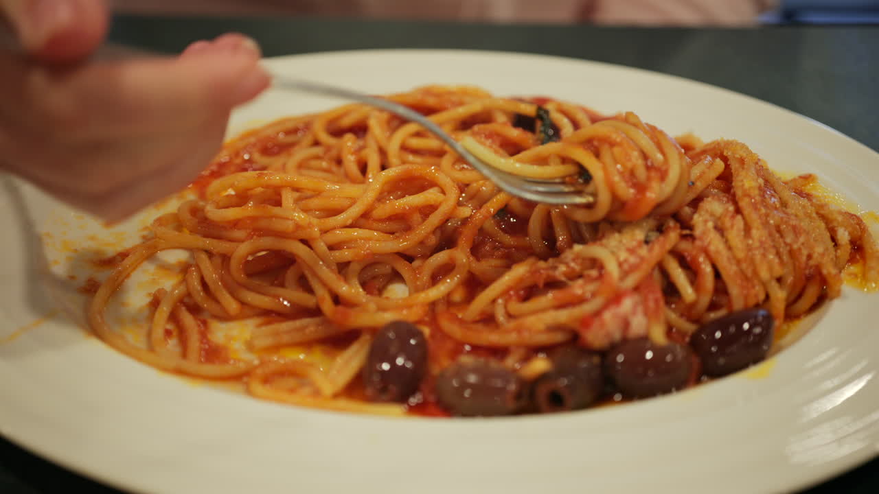 Primo piano di una persona che avvolge spaghetti alla puttanesca con una forchetta, mostrando un ricco sugo di pomodoro, olive e formaggio grattugiato