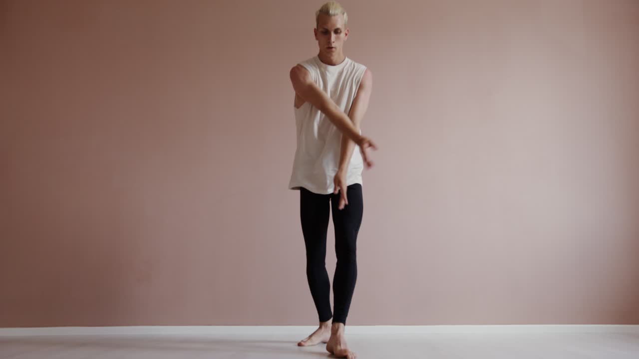 A young man in a white tank top and black leggings dances in a studio