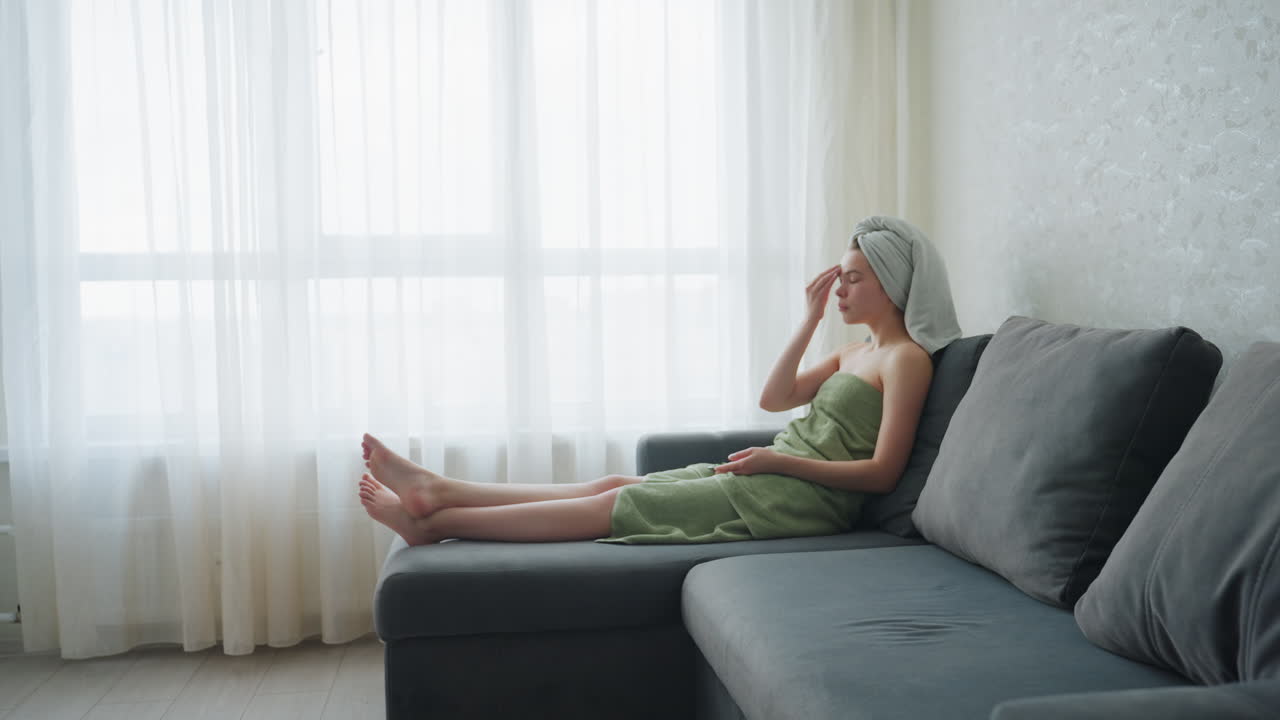Woman wrapped in towel sits on couch with legs stretched out, gently rubbing cucumber slice on forehead in calm room with soft natural light, enjoying relaxing skincare and self-care moment