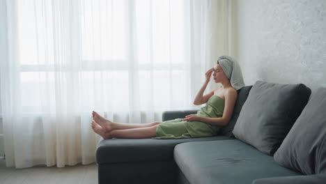 woman wrapped in towel sits on couch with legs stretched out, gently rubbing cucumber slice on forehead in calm room with soft natural light, enjoying relaxing skincare and self-care moment