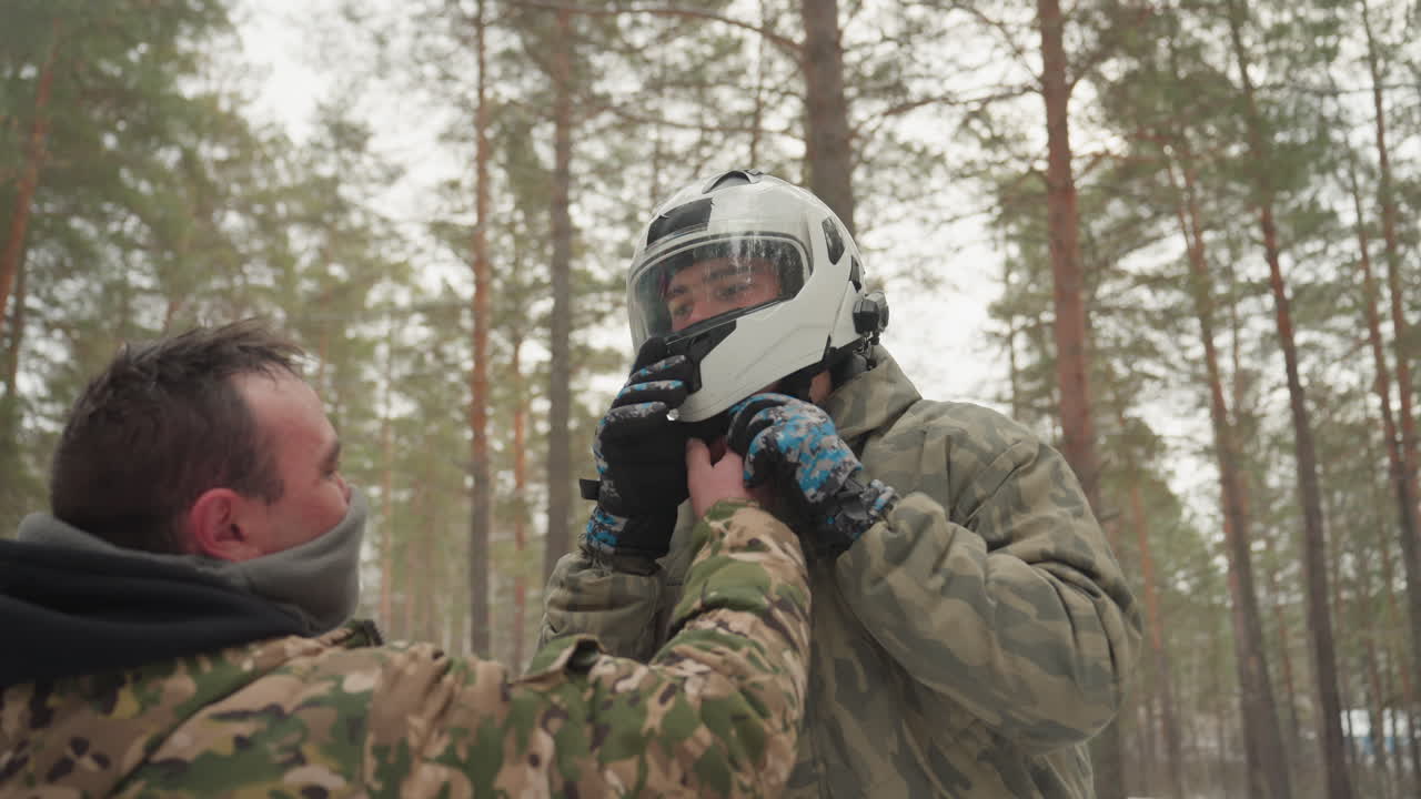Snow forest atv helmet being adjusted and buckled by helper in camouflage, close up on chinstrap and headgear. Twoperson prep scene shows training, inspection, and teamwork before offroad ride careful
