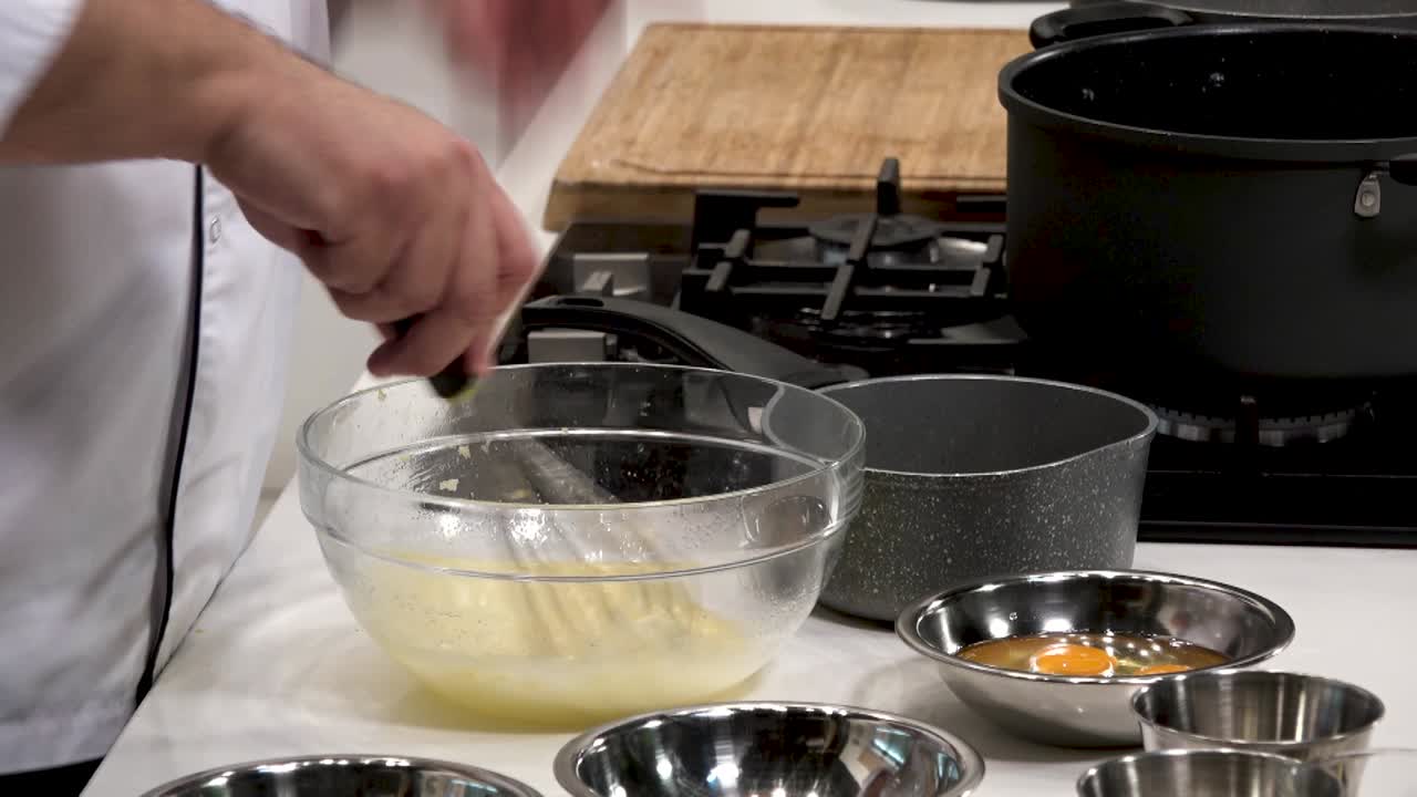I ett modernt kök vispar en kock skickligt ihop ägg och andra ingredienser i en klar skål. Omkringliggande skålar innehåller ytterligare ingredienser, redo att användas.