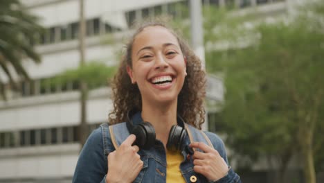 Retrato-De-Una-Mujer-Birracial-Feliz-En-La-Ciudad,-Usando-Audífonos-Y-Sonriendo