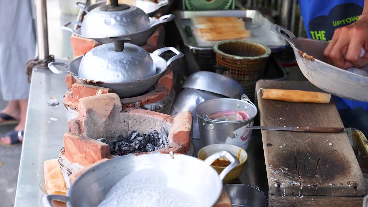 Hands prepare coconut snacks on clay stoves in a bustling Phuket market. Bright lighting highlights traditional cooking techniques