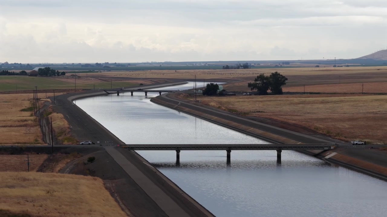 Breites Beton-Aquädukt mit Straßenbrücke, das sich durch weite, flache landwirtschaftliche Felder und trockenes goldenes Ackerland in Richtung einer entfernten Baumgrenze unter bewölktem Himmel erstreckt. Kalifornien, USA. Luftaufnahme Weitwinkel