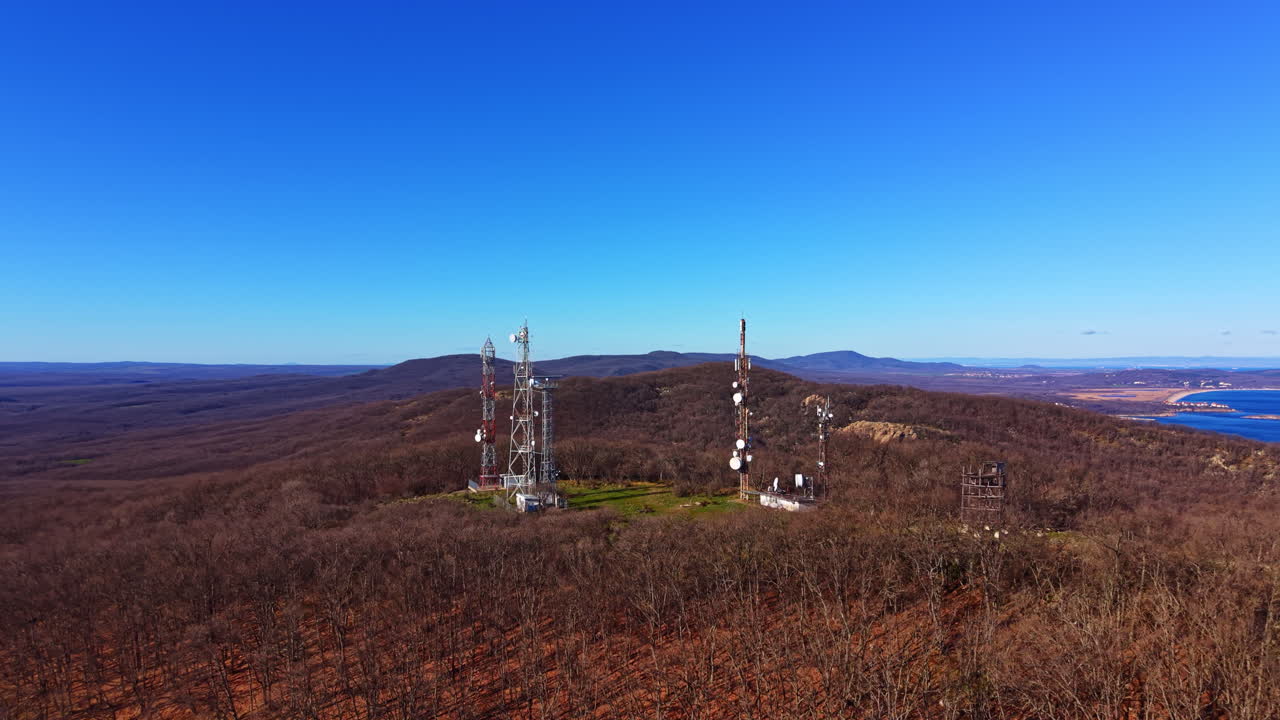 Communication towers stand on a mountain top overlooking a valley and a river under a clear blue sky during daytime