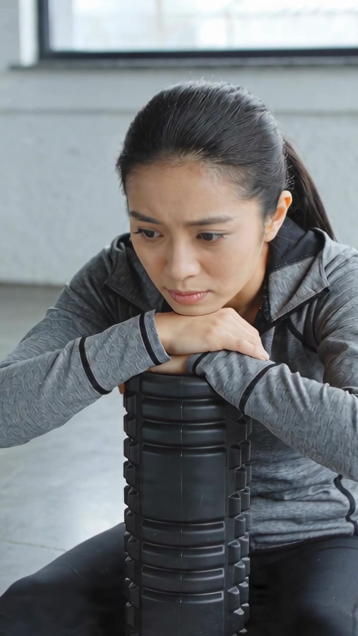 Vertical video: Leaning woman in grey jacket on foam roller at gym, recovering after workout