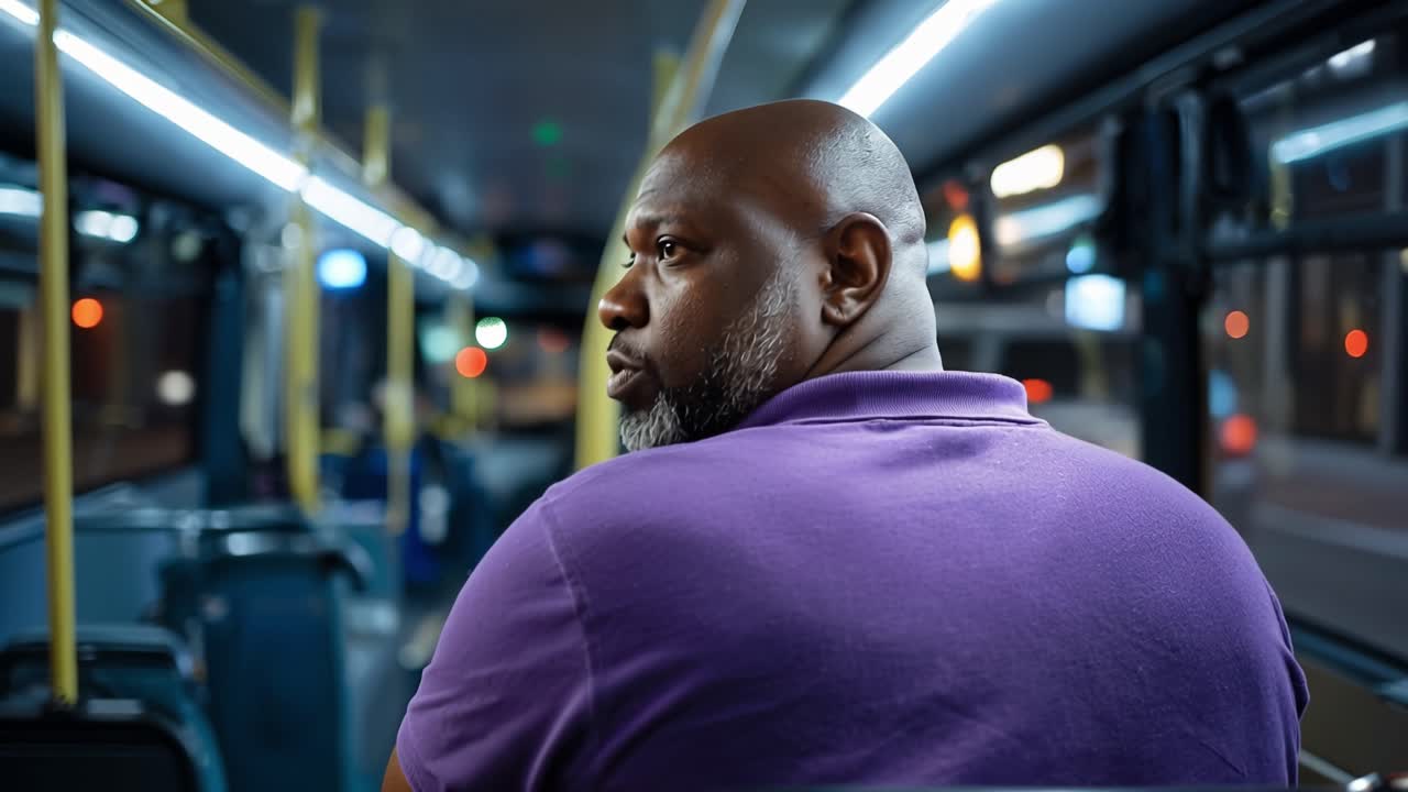 Man sits alone on a bus at night. He stares forward, quiet and thoughtful. Lights glow softly along the bus's ceiling. He wears a purple shirt, calm and still. The city blurs past, unseen and distant