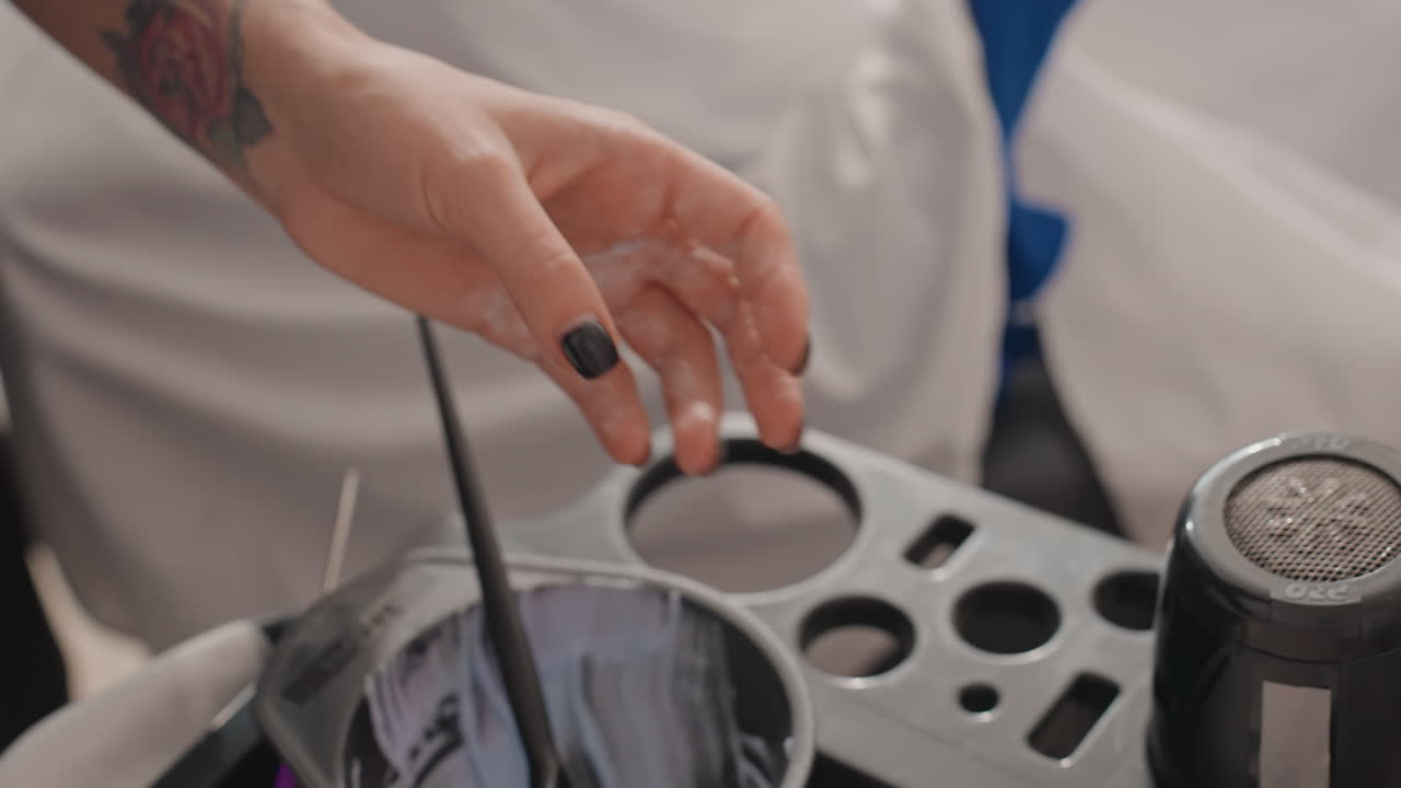 Hænder i en salon, der lægger foliestriber og forbereder farveskål med applikatorbørste, nærbillede af tatoveret håndled og sort manicure, metalliske folieark foldet til balayage-teknik, blandekopper