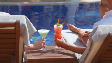 unrecognizable couple enjoying refreshing drinks lying on sunbeds on poolside