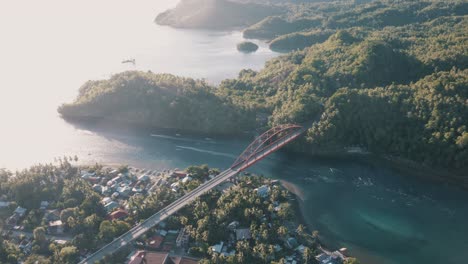 Vehículos-Y-Motocicletas-Que-Pasan-Por-El-Puente-De-Metal-Dorado-Que-Conecta-El-Pequeño-Pueblo-De-Pescadores-Y-Las-Exuberantes-Montañas-Con-Vista-Al-Océano-Tropical-En-Leyte,-Filipinas