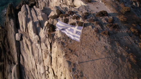 aerial view of greek national flag waving on cliff above aegean sea, drone shot