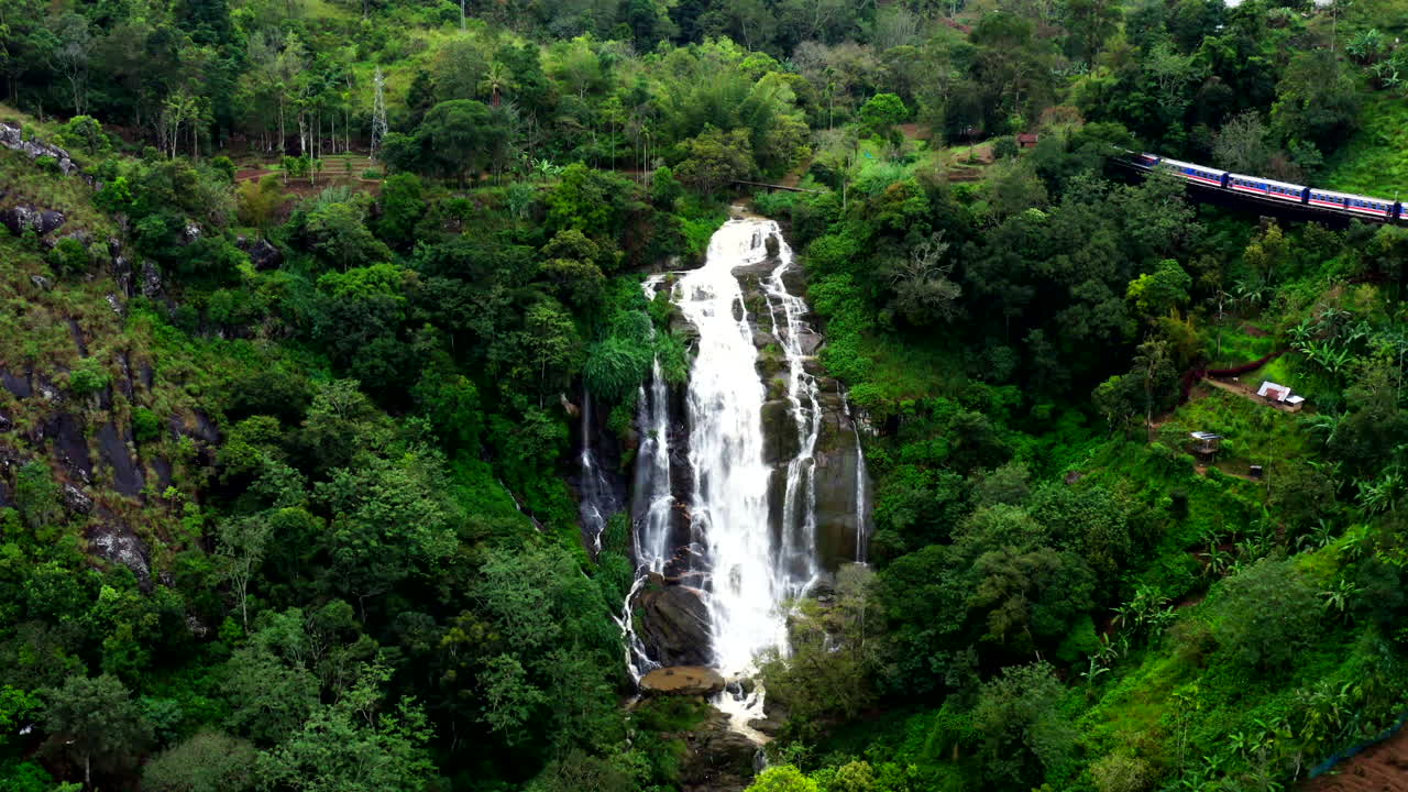 Drone shot of a beautiful waterfall near Ella, Sri Lanka, with a passing train crossing a bridge in the lush tropical landscape