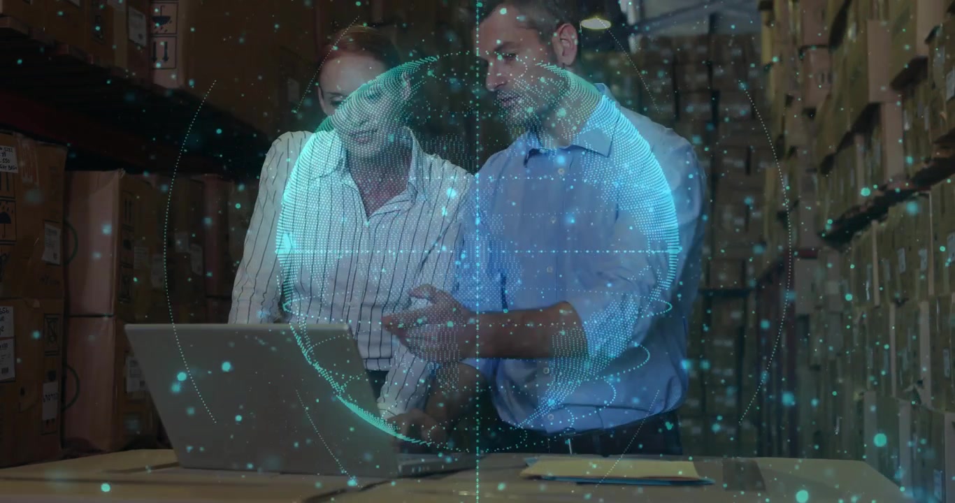 female and male colleagues leaning over laptop in warehouse, showcasing logistics data hologram