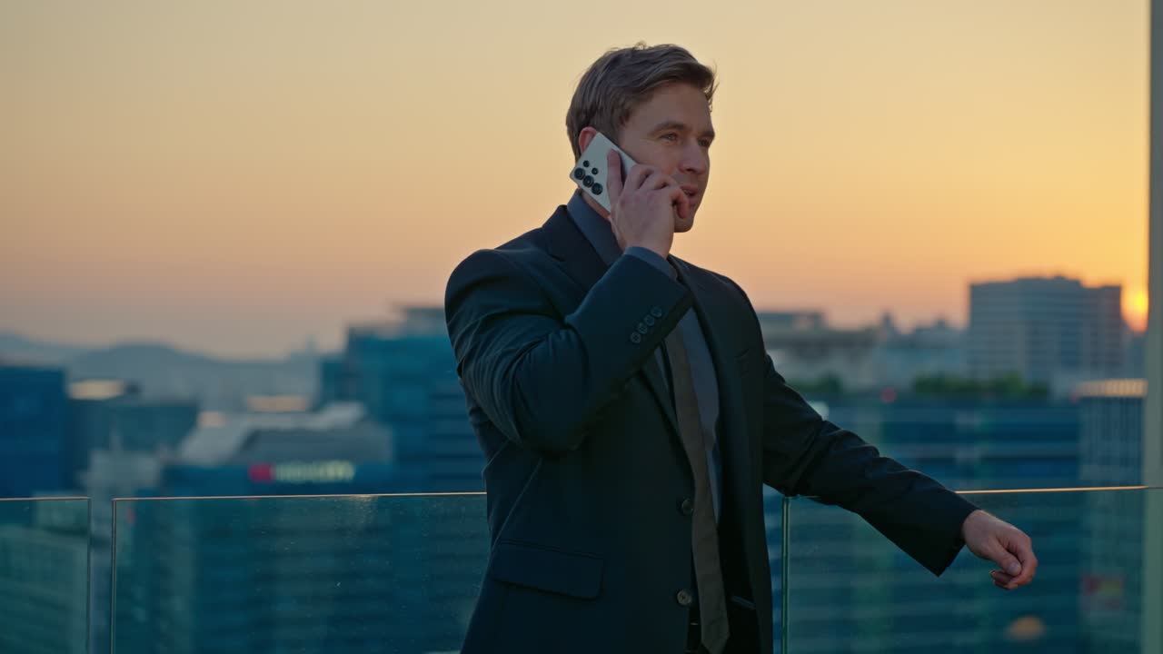 A happy Caucasian businessman with a toothy smile talks on his smartphone in slow motion, with the Seoul cityscape and sunset in the background from a rooftop