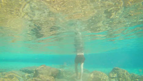 young man walking in blue clear under water of waterfall with many rocks from flat angle