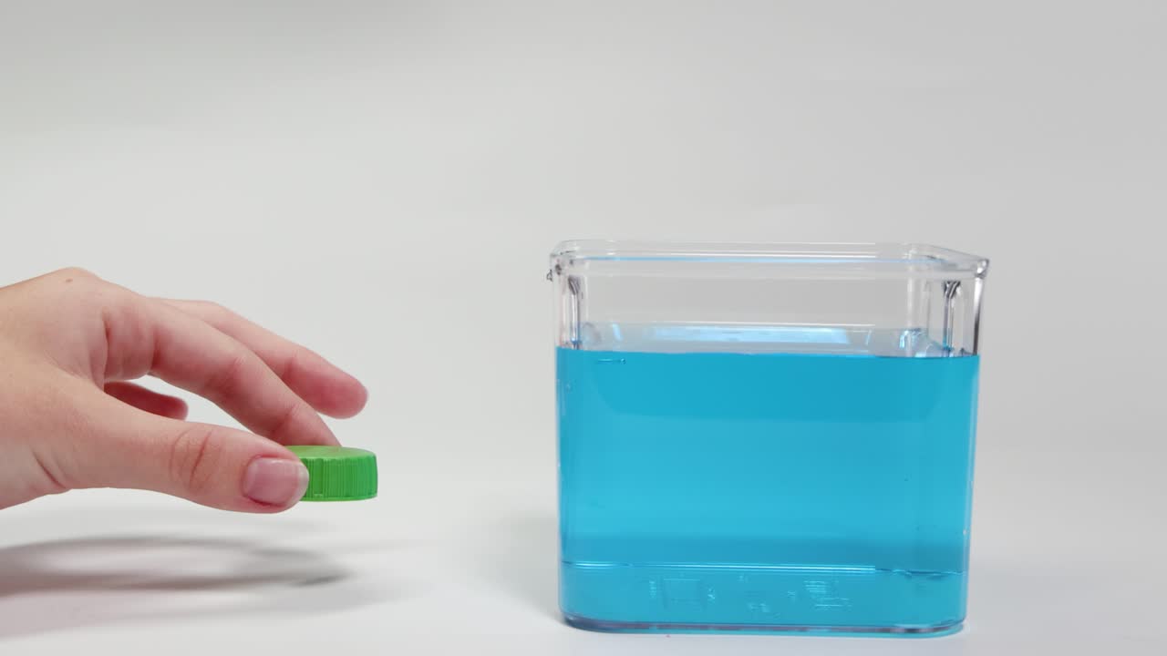 A hand places a green plastic cap in blue water, demonstrating buoyancy in a clear container