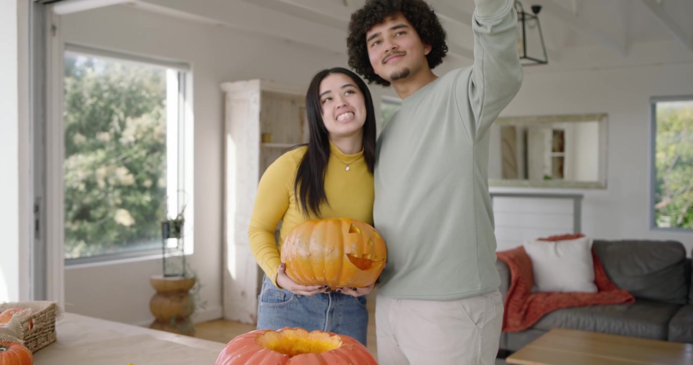 Coppia che finisce di intagliare su un tavolo di quercia, donna con un top giallo in posa con il telefono per fotografare la zucca
