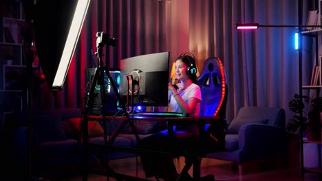 asian girl streamer playing game over network on personal computer, waving hand talking to the participants on the microphone.  live stream video game, desk illuminated by rgb led strip light