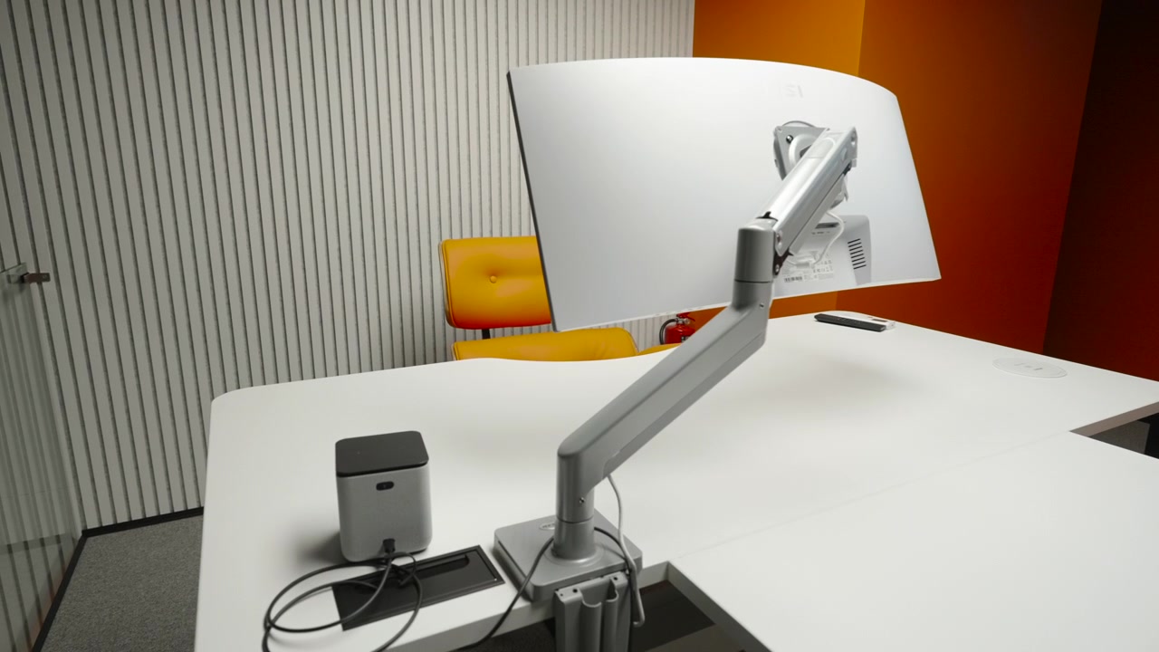 Bright contemporary workspace with white desks, metal monitor arms, striped acoustic panels, an orange accent wall, and a clean gray carpeted floor under soft