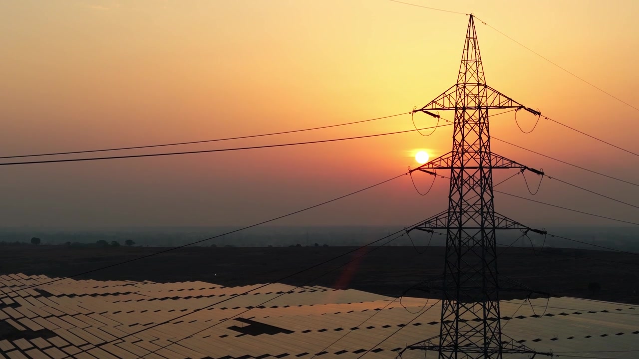 Drone ascending over a vast solar power plant with high voltage transmission tower at sunset, showcasing renewable energy infrastructure, electricity generation, and sustainable power.