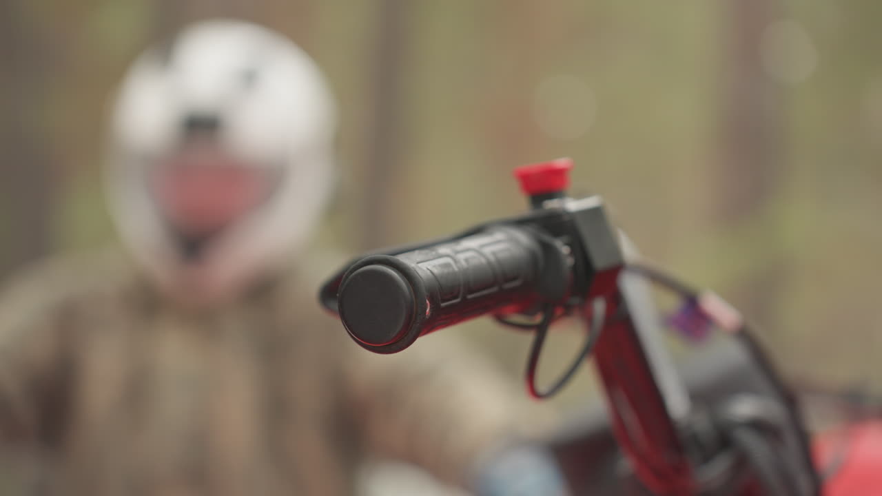 Nærbillede af ATV-styr i skov, hjelmklædt kører justerer gas og greb, synligt camo-jakkeærme, rød stopknap som fokuspunkt, sløret fyrretræbaggrund, spændt forventning før terrænkørsel.