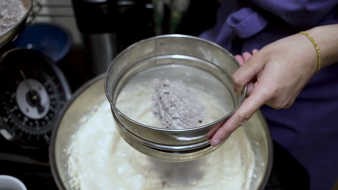 Close up of a pastry chef sifting a mix of flour and cocoa powder through a sieve into a bowl, preparing ingredients for baking a cake