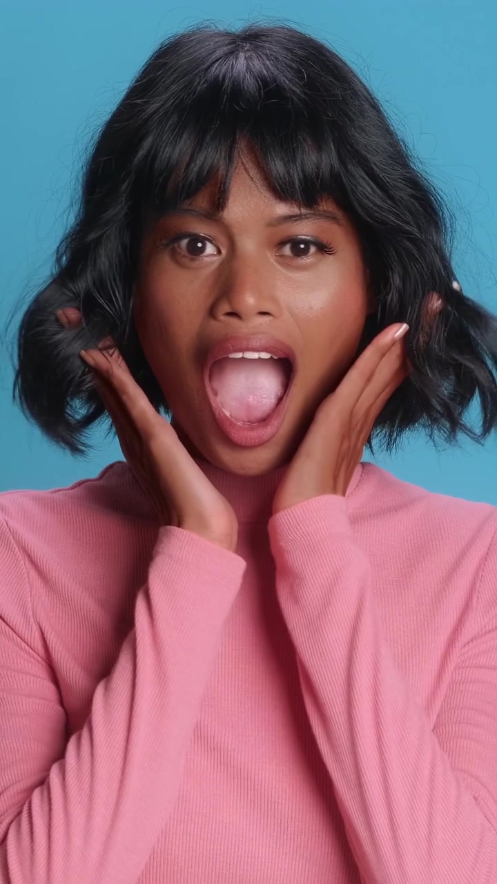 Surprised asian woman holding cheeks in shock and excitement on blue studio