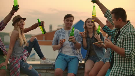 three girls and three boys in plaid shirts drinks beer from green bottels on the party with friends on the roof at the sunset.they are sitting together eat hot pizza after in summer everning. a man says a toast to his friend's birthday.