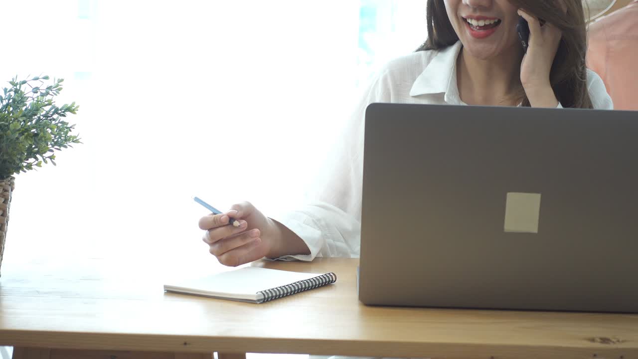 Bella giovane donna asiatica sorridente che lavora al laptop mentre è seduta in salotto a casa. Donna d'affari asiatica che usa il telefono per lavoro nel suo home office. Si gode il tempo a casa