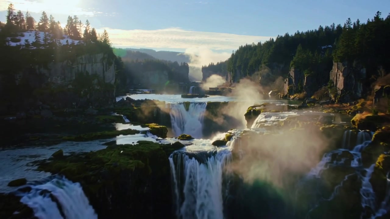 Gliding drone camera moving from wide river valley toward multi-tier waterfall, revealing mist