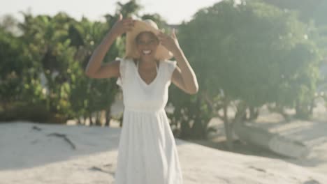 Retrato-De-Una-Mujer-Birracial-Feliz-Con-Vestido-De-Verano-Y-Sombrero-Bailando-En-La-Playa-Soleada,-Cámara-Lenta