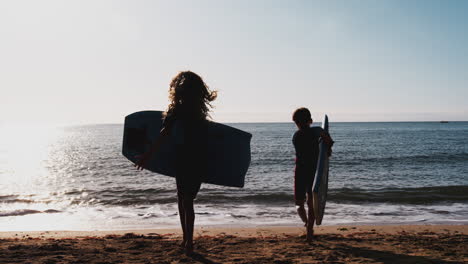 Rückansicht-Von-Zwei-Silhouettierten-Kindern-In-Neoprenanzügen,-Die-Im-Strandurlaub-Mit-Bodyboards-Ins-Meer-Laufen