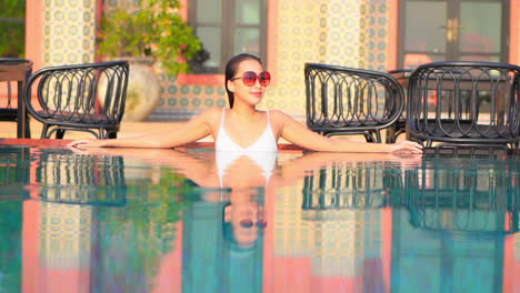 Una-Mujer-De-Lujo-Con-Traje-De-Baño-Blanco-Y-Gafas-De-Sol-Rojas-Apoyada-En-El-Borde-De-La-Piscina-Del-Hotel-Al-Atardecer